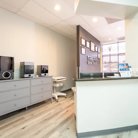 Bright dental office reception area with gray cabinets, equipment, and Trophy Smile Studio sign on the wall.