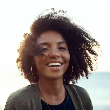 Smiling woman with curly hair against a beach sunset background, exuding warmth and happiness.