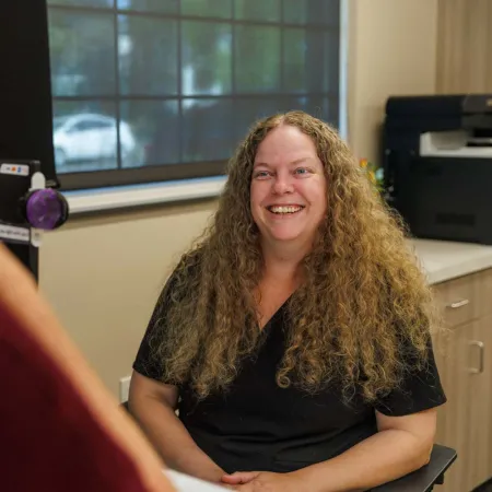 Smiling woman with curly hair in black shirt talking to another person in office setting with window behind.