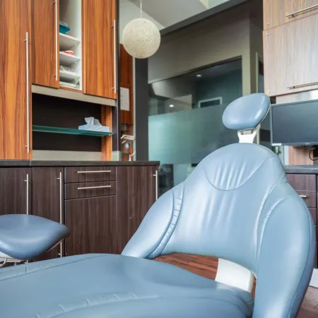 Modern dental office with a blue patient chair, wooden cabinets, and computer monitor in the background.