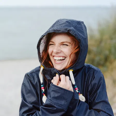 A woman in a rain jacket smiles joyfully by the beach on a rainy day.