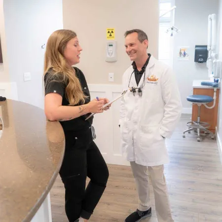 Female medical staff with clipboard talking to male doctor in white coat at clinic reception area.