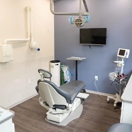 Modern dental clinic room with dental chair, equipment, monitor, and clean bright interior with wood flooring.