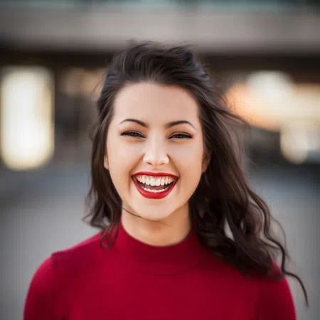 Smiling woman with red lipstick and red top, curly hair blowing in the wind outdoors with blurred background.