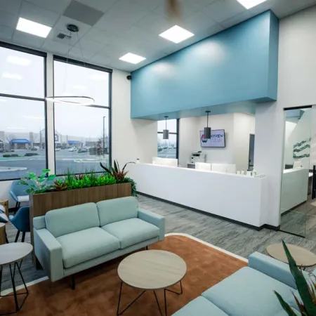 Modern office lobby with light blue couches, wood tables, indoor plants, large windows, and white reception desk.
