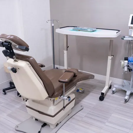 Medical examination room with adjustable brown patient chair, rolling tray table, and medical equipment on stand.