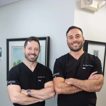 Two male doctors in black scrubs posing with arms crossed, smiling in a medical office setting.