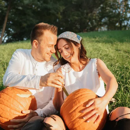 Young couple carving pumpkins outdoors on a sunny day sitting on grass with greenery in the background.