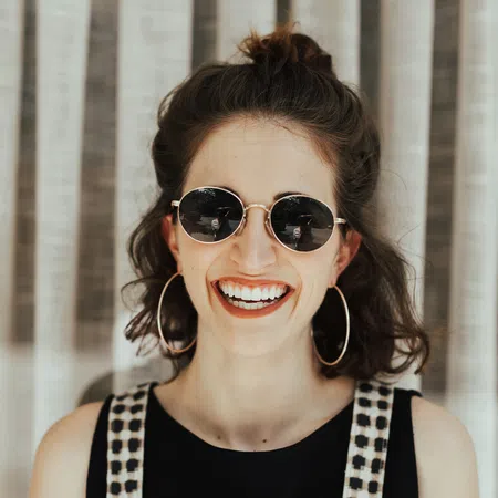 Young woman wearing sunglasses and hoop earrings, smiling in front of a curtain backdrop.