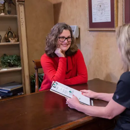 Two women discussing documents at an office desk amid framed certificates on the wall.