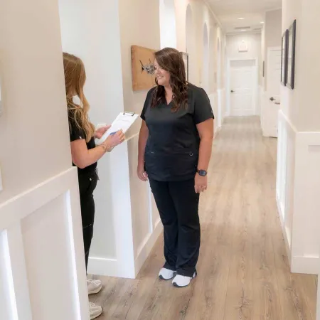 Two healthcare workers in black scrubs conversing in a bright medical hallway with white walls and wooden flooring.