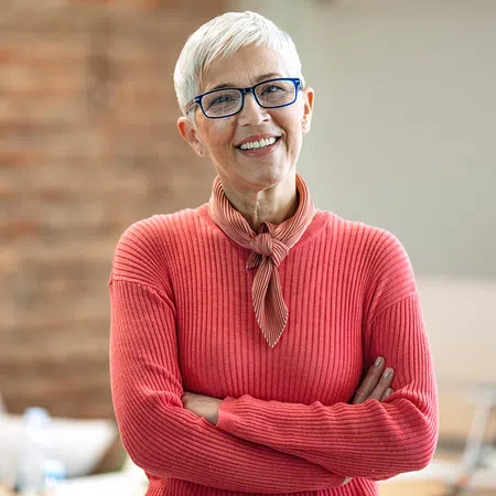 Smiling older woman in a sweater, arms crossed, standing in an office setting with a blurred background.