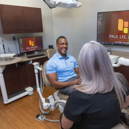 Dentist and patient discussing dental care in a modern clinic with screens displaying Paul Lee DDS logo.