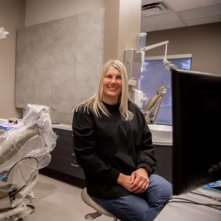 Smiling female dentist in black scrubs sits in a modern dental office with chair and dental X-ray screen.