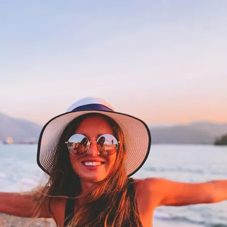 Smiling woman in hat and sunglasses enjoying sunset on rocky beach with arms outstretched by the sea.