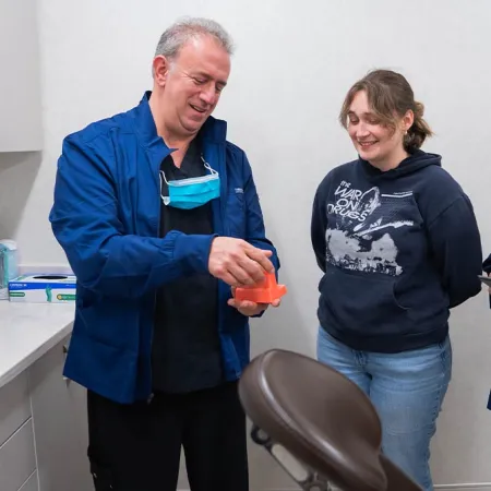 Dentist explains dental model to female patient with dental assistant holding tablet in clinic room.
