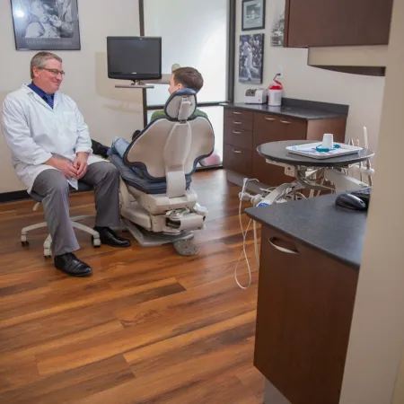 Dentist in white coat talking with patient seated in dental chair in modern clinic room with wood floor.