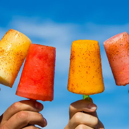 Four hands holding colorful fruit popsicles with chili powder against a clear blue sky background