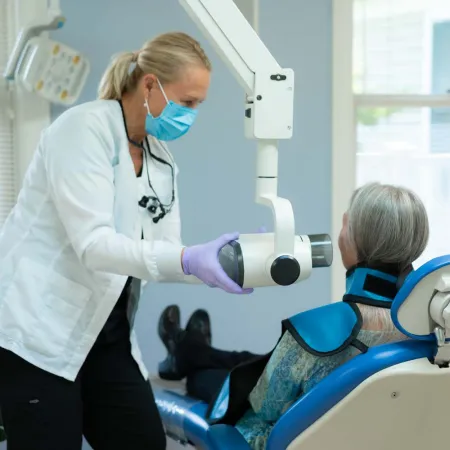 Dentist wearing mask prepares X-ray machine for elderly female patient seated in dental chair