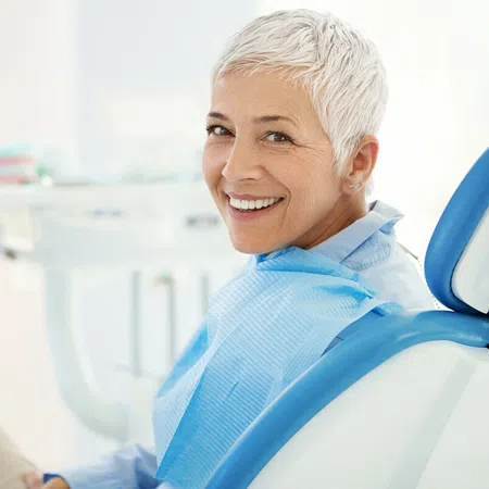 Smiling older woman in a dental office, seated in a chair with dental tools visible.