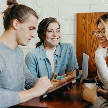 Group of young adults collaborating and smiling while using a tablet and laptop in a cafe setting.