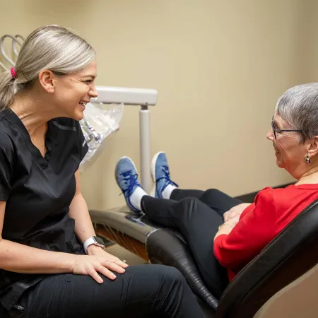 Female dentist smiling and talking with senior female patient sitting in dental chair in clinic room.