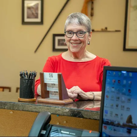 Smiling older woman in red shirt behind counter talking to a customer in an office setting with computer and pens.