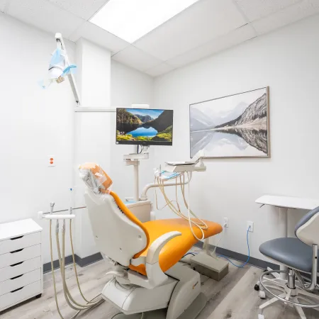 Bright dental clinic room with orange patient chair, dental equipment, monitor, and nature artwork on white walls.