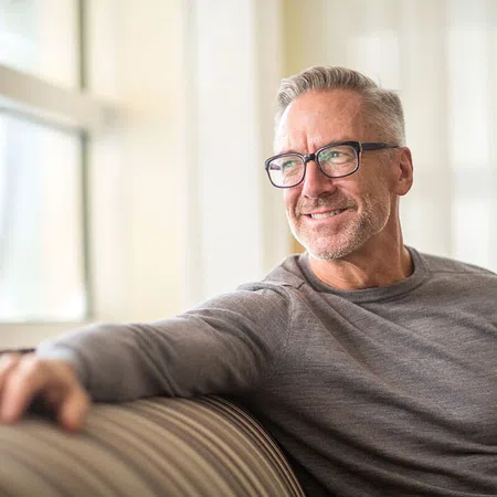 Smiling middle-aged man in glasses relaxing on a sofa by a window.