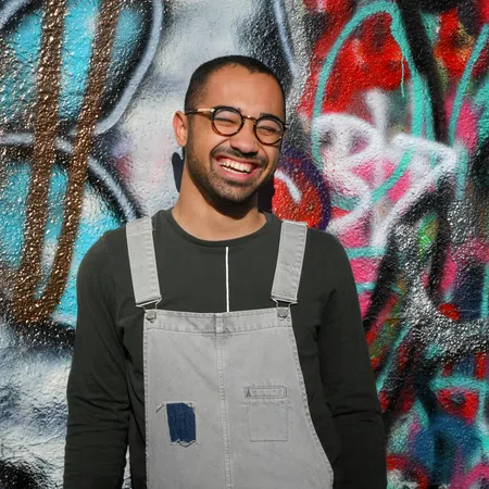 Smiling man with glasses wearing grey overalls and holding a black cap stands against colorful graffiti wall.