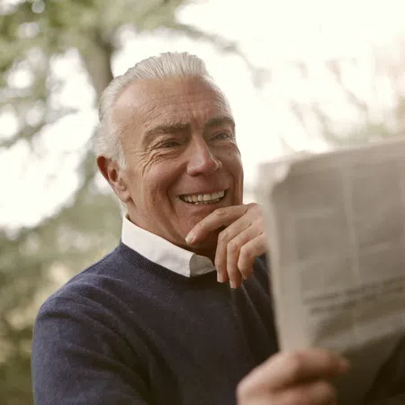 Smiling senior man reading a newspaper outdoors with blurred natural background in daylight.