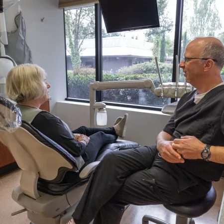 Dentist and patient sitting facing each other in a bright dental office with large windows and modern equipment