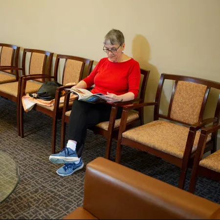 Elderly woman in red shirt reading magazine in a waiting room with chairs and glass table.
