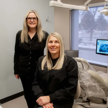 Two female dentists in black uniforms posing in a modern dental office with equipment and X-ray on screen.