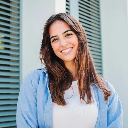 Smiling young woman with long hair wearing a blue shirt, standing in front of green shutters.