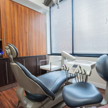 Modern dental office with black dental chair, treatment equipment, wooden cabinets, and large window with blinds.