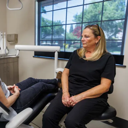 Patient seated in dental chair talking with female dental professional in modern clinic with large windows.