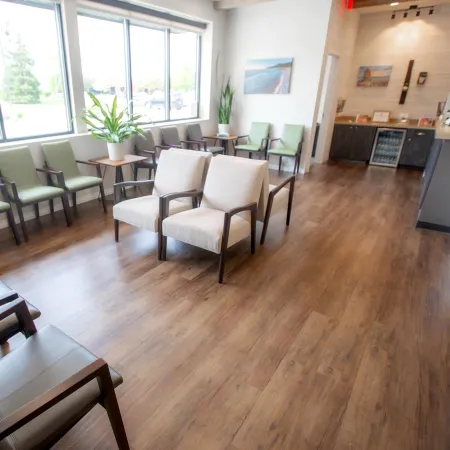 Bright waiting room with wood flooring, beige and green chairs, large windows, plants, and a refreshment area.