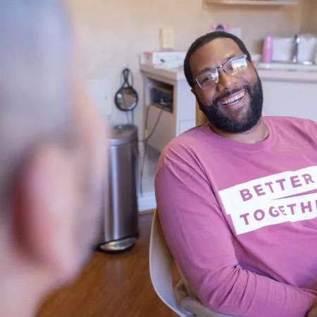 Smiling man in glasses wearing a pink sweatshirt that says Better Together talking indoors with another person.