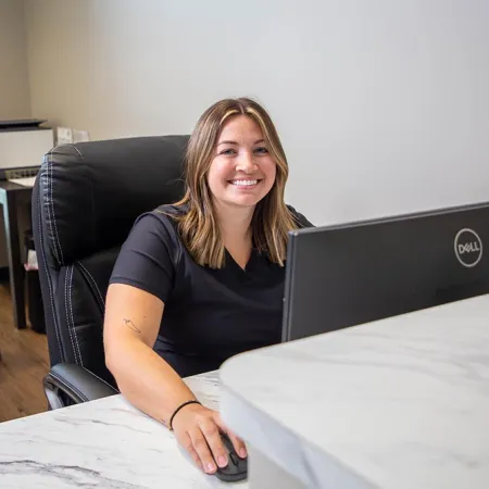 Smiling woman working at a desk with computer in a modern office space with another woman in background.