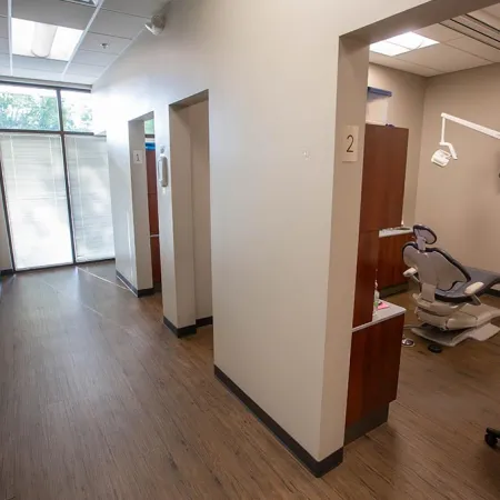 Modern dental clinic hallway with treatment rooms featuring dental chairs, TV monitors, and wood flooring.