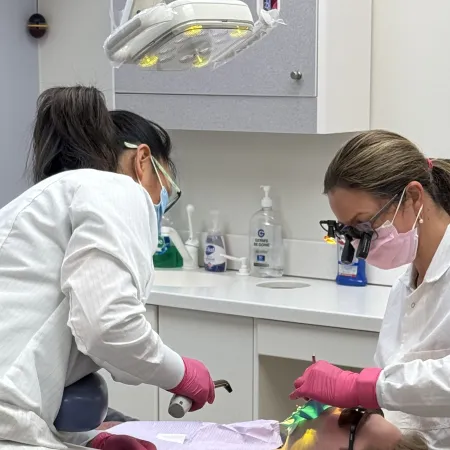 Two dentists wearing masks and gloves perform a dental procedure on a patient in a clinic room.