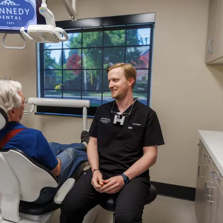 Dentist converses with elderly patient in modern Kennedy Dental office with large window and dental chair.