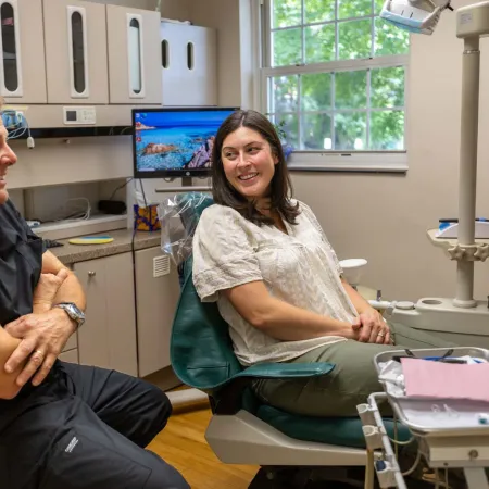 Dentist and patient smiling and talking in a bright dental office with equipment and monitors.