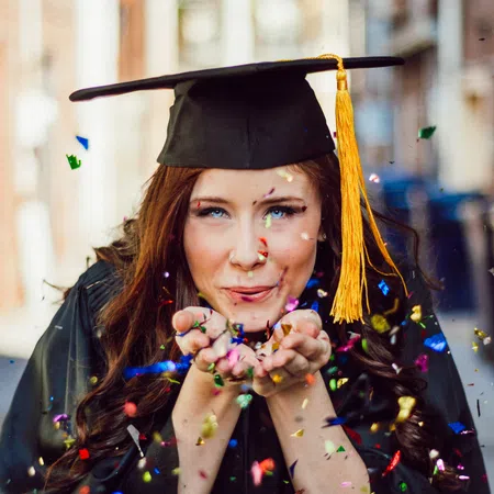 Young female graduate wearing a cap and gown blowing colorful confetti in celebration outdoors.