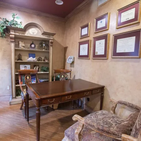 Elegant office with wooden desk, framed certificates, patterned chairs, and a decorative bookcase with plants.