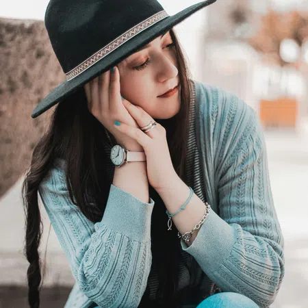 Young woman wearing a black hat and gray sweater, resting her face on her hands with closed eyes outdoors.