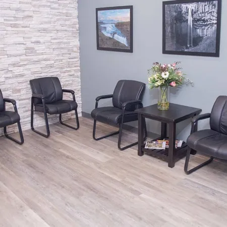 Modern waiting room with four black leather chairs, a wooden table with flowers, and framed wall art.