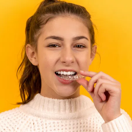 Young woman in white sweater inserting a clear dental aligner against a bright yellow background.