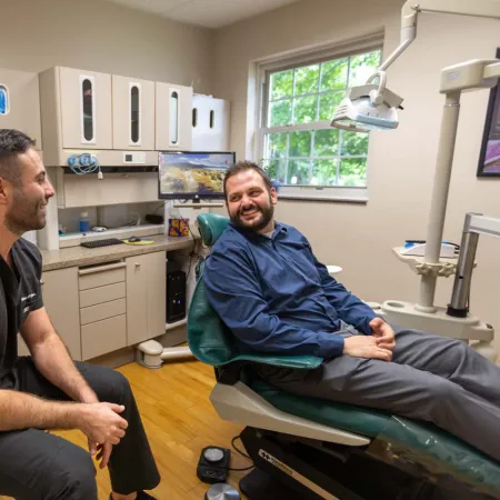 Dentist in black scrubs consulting a smiling male patient sitting in a dental chair in a bright clinic room.